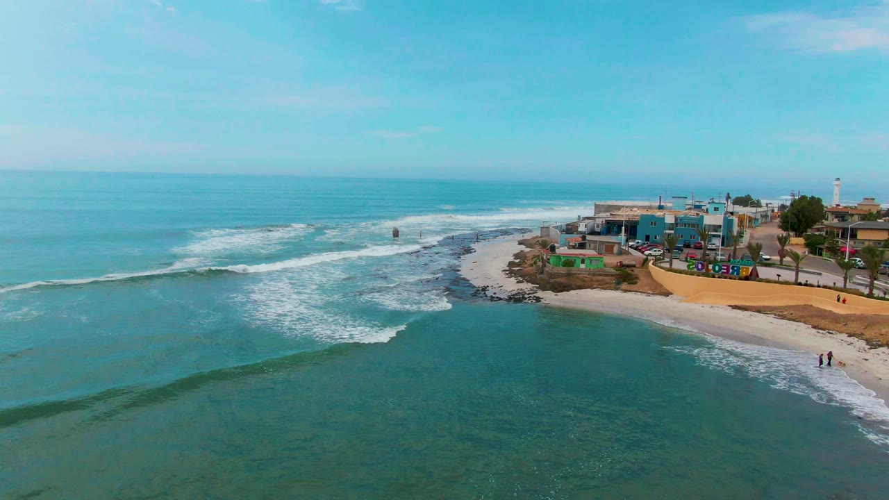Punta Abreojos coastline seen from above, with turquoise water filling the foreground, surf wrapping around a rocky point, buildings on the right, and tiny people near the lower beach.