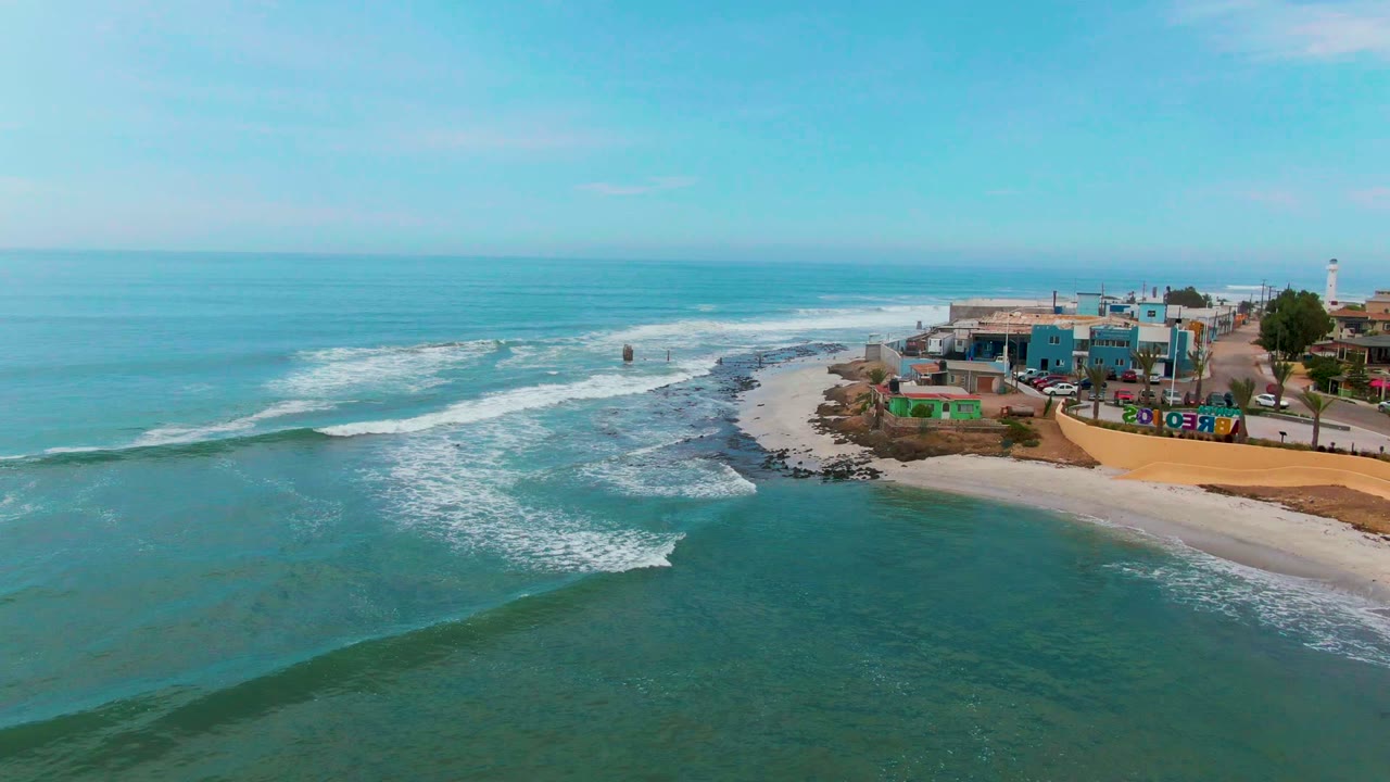 Ocean waves break toward a beach beside compact coastal buildings.