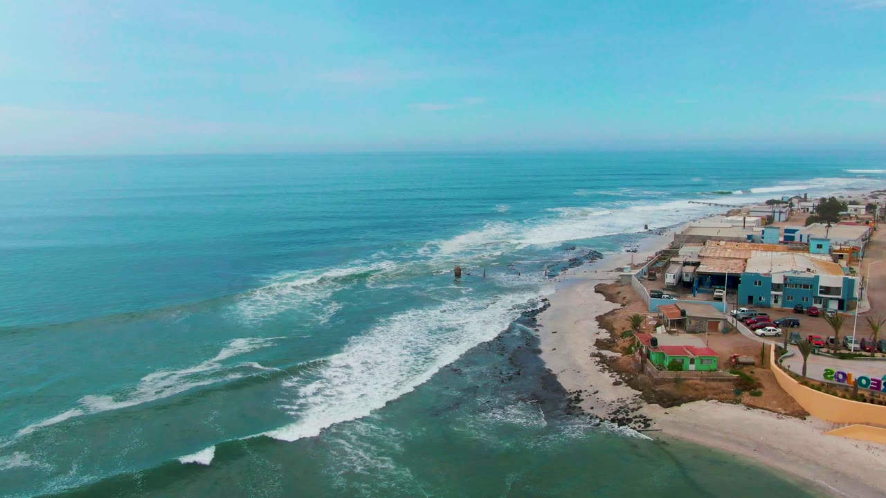 Aerial view of Punta Abreojos, Baja California Sur with turquoise ocean, white surf, dark shoreline rocks, sandy beach, coastal buildings, parked cars, and no visible people.