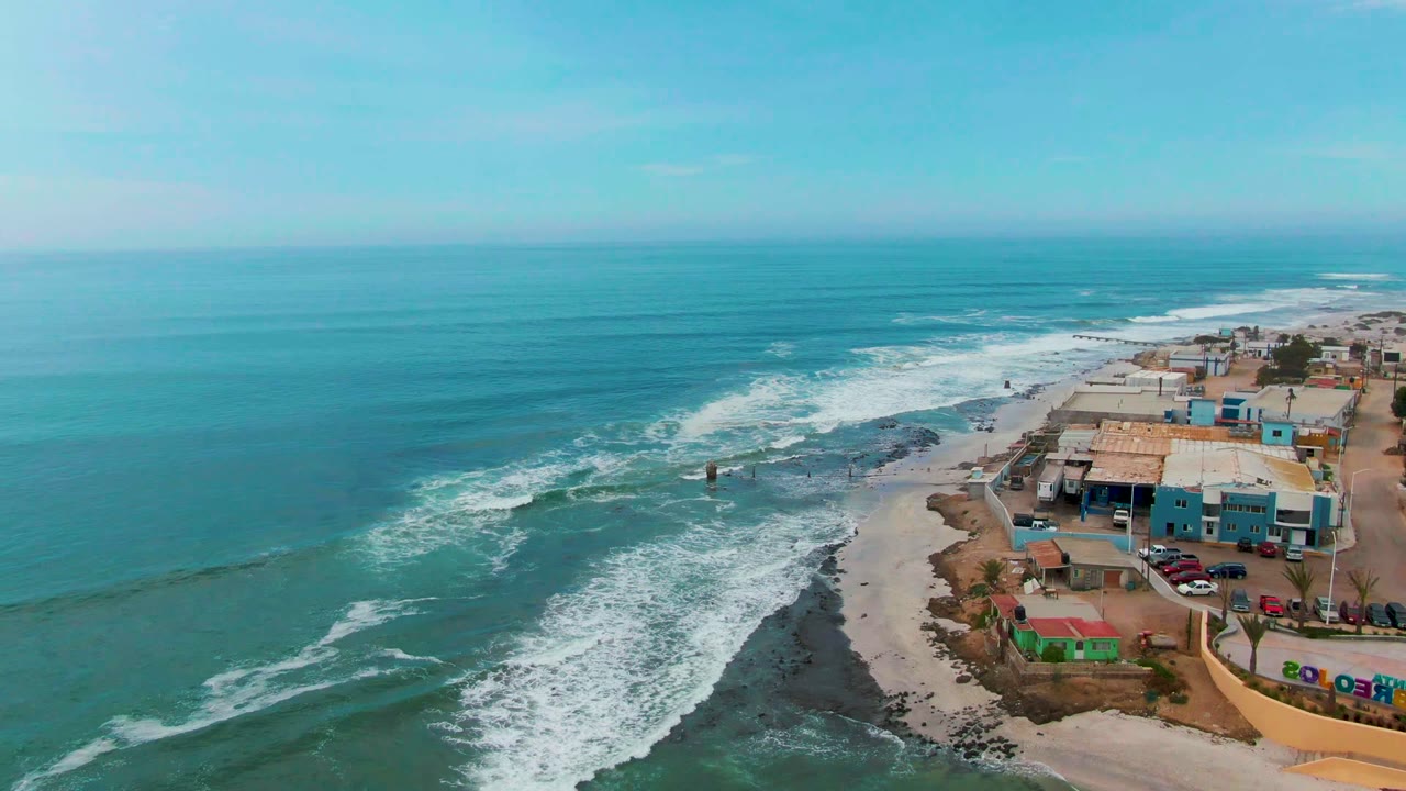 High coastal view in Punta Abreojos with broad blue ocean, a diagonal band of breaking waves, dark reef-like rocks, sandy beach, low buildings, cars, and no visible people.