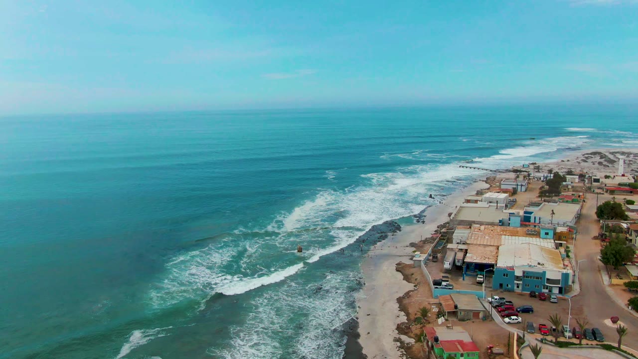 A high aerial frame over Punta Abreojos shows turquoise ocean on the left, white surf breaking along the shore, and low beachfront buildings with parked cars on the right.