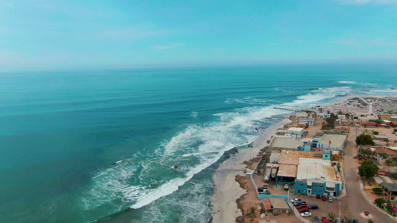 At 0:02, white wave bands curve diagonally toward the beach while blue and tan beachfront structures and a few cars line the right edge of the Punta Abreojos coast.