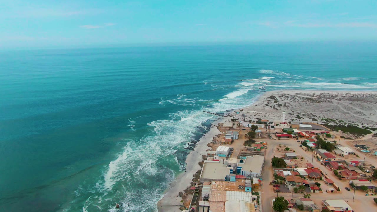 Aerial view over Punta Abreojos, Mexico, with turquoise ocean on the left, white surf breaking along rocks, sandy land and low buildings on the right, and no visible people.