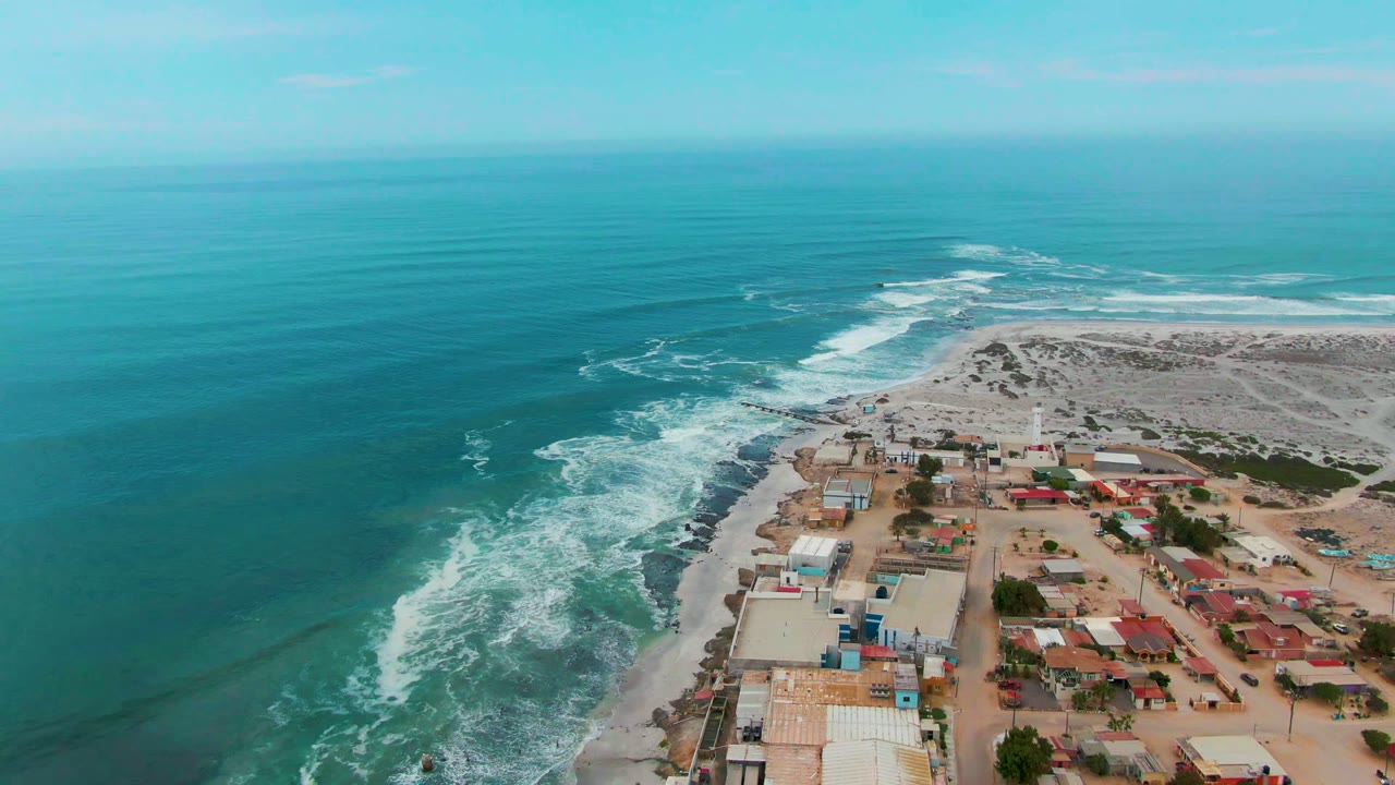 Aerial coastline with white surf curling along rocks and buildings on sandy land.