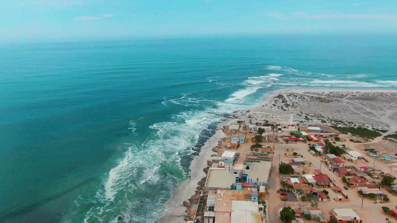 Aerial frame over Punta Abreojos with darker ocean swell at left, bright foam along the rocks, pale dunes in back, and red-roofed and light-roofed buildings at lower right, with no visible people.