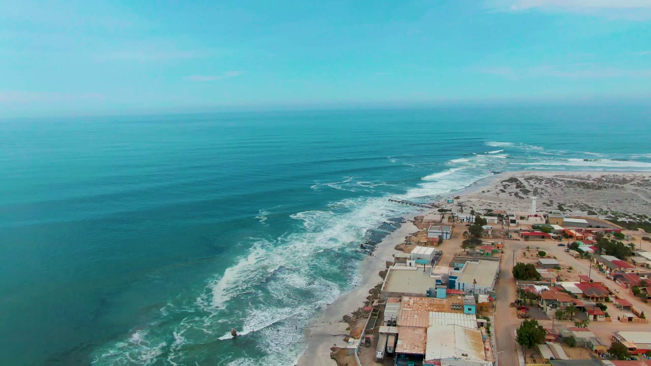 Aerial frame over Punta Abreojos, Baja California Sur showing turquoise ocean, white surf, beachside buildings, pale dunes, and no visible people at the start of the clip.