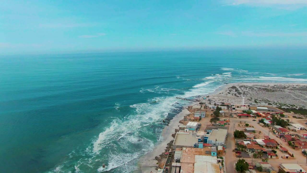 Aerial frame over Punta Abreojos showing the town edge close to the beach, white water along the shore, calm blue-green sea beyond, and no visible people.