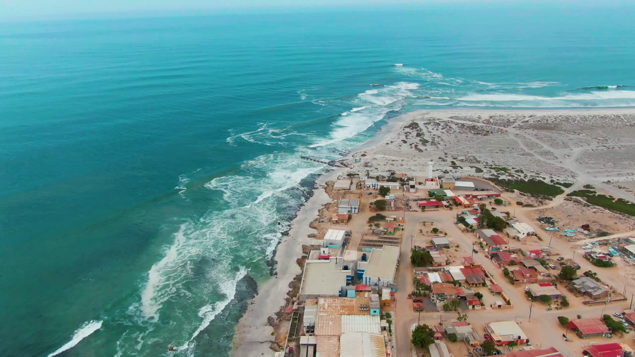 Opening aerial frame over Punta Abreojos, Baja California Sur, with turquoise water on the left, white surf along the shore, low roofs on sandy terrain, and no visible people.