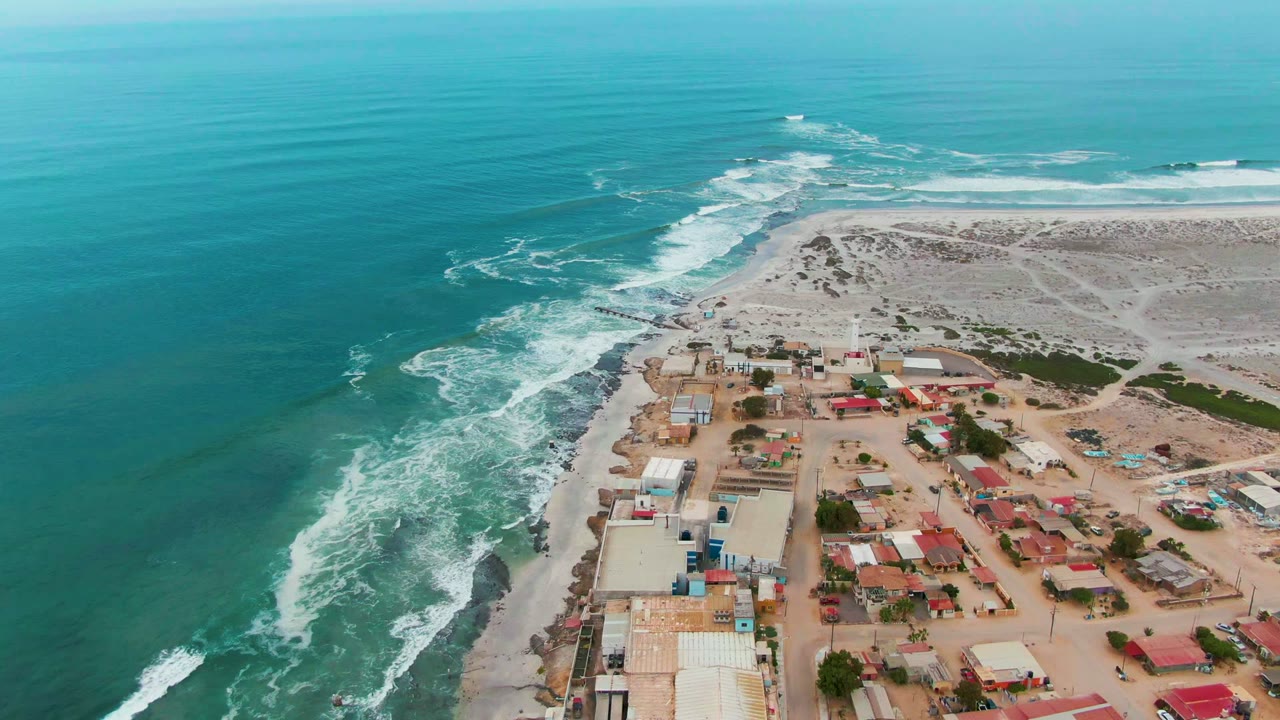 Drone view of white surf wrapping around a sandy point beside low coastal buildings.