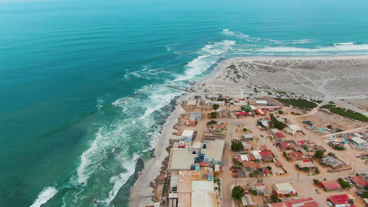 Mid-clip aerial frame over Punta Abreojos showing the ocean pushed farther left in frame, clustered red and gray roofs on the lower right, pale shore sand, and no visible people.