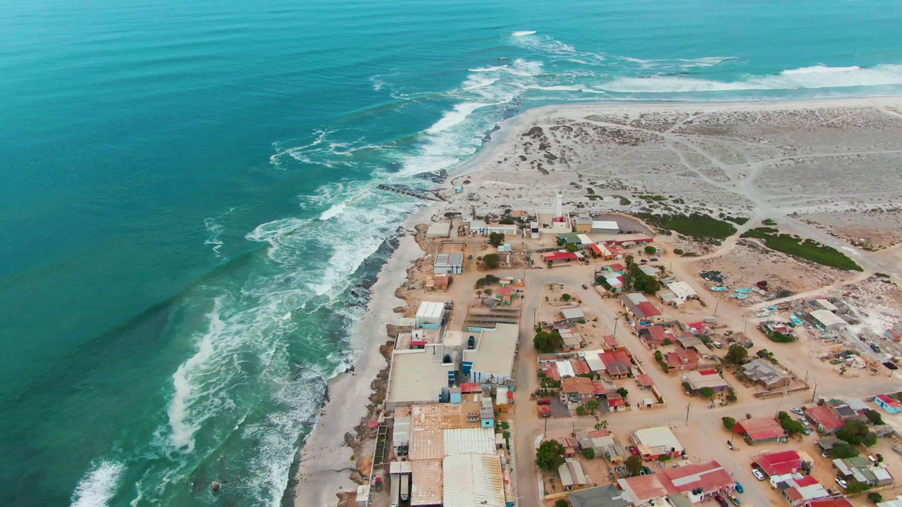 Opening aerial frame over Punta Abreojos, Baja California Sur, with turquoise ocean on the left, white waves along the shore, low buildings on sand, pale dunes beyond, and no visible people.