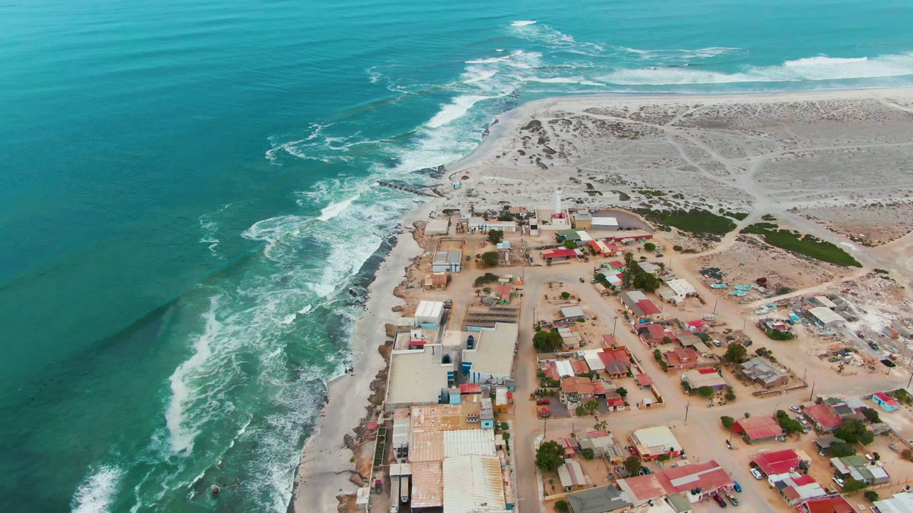 Turquoise water and breaking waves beside low buildings on a sandy shore.