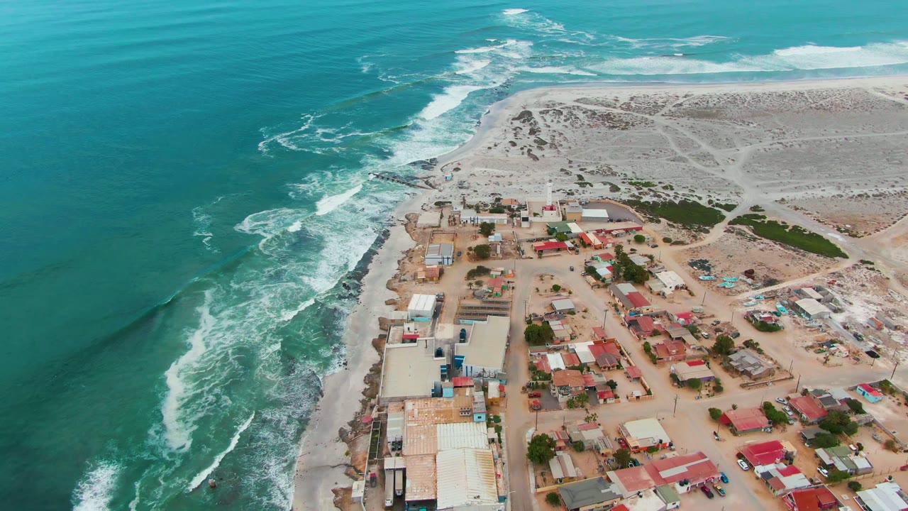Punta Abreojos appears from above with broad turquoise water to the left, foamy waves curving toward shore, compact village buildings below, sandy tracks across the dunes, and no visible people.