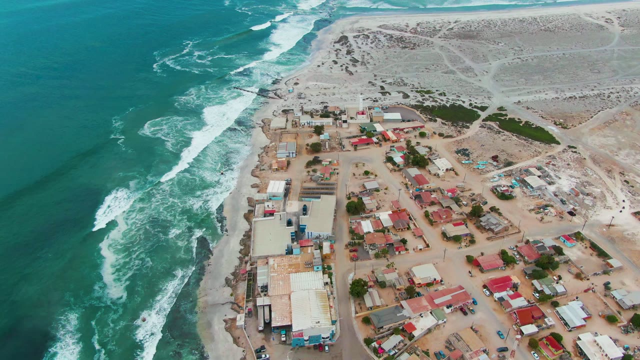 Aerial view over Punta Abreojos, Baja California Sur, with turquoise surf on the left, red-roofed buildings near the beach, pale dunes inland, and no visible people.