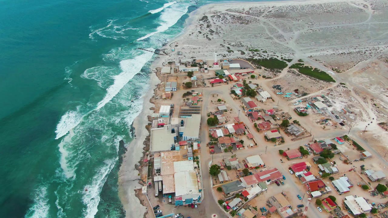 High drone frame of Punta Abreojos showing the surf line closer to the settlement edge, clustered small buildings, sandy lots, dunes, and no visible people.