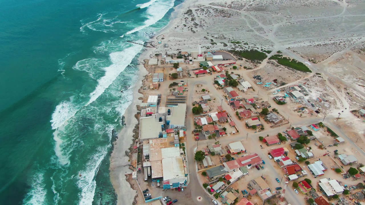 Aerial frame over Punta Abreojos with a broad band of turquoise water, foamy waves, a compact sandy-grid settlement, scattered vehicles or small objects, dunes, and no visible people.