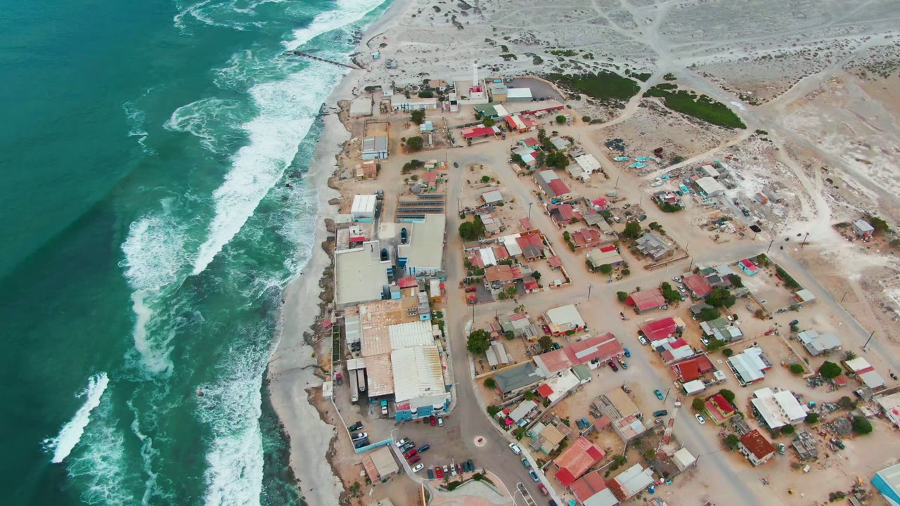 Aerial frame over Punta Abreojos, Baja California Sur, with blue-green ocean on the left, white surf along the shore, low buildings and roads on the right, and no visible people.