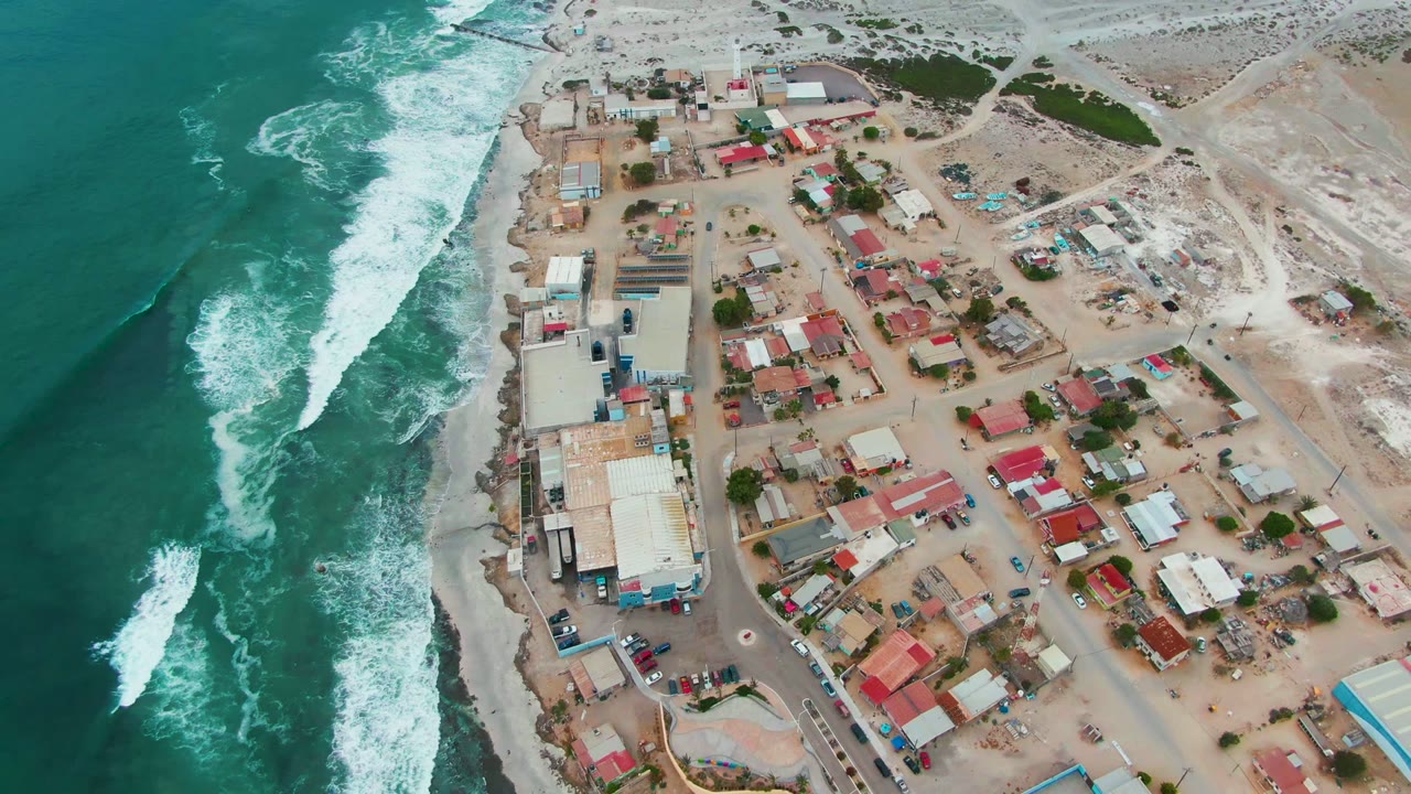High aerial frame of Punta Abreojos showing surf breaking in staggered white lines, a sandy town grid with red roofs and blue-roofed structures, vehicles, and no visible people.