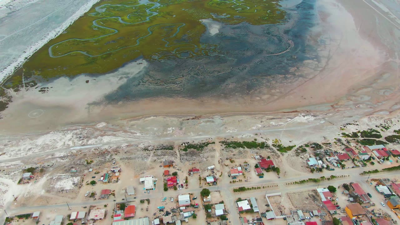 Aerial frame over Punta Abreojos, Baja California Sur, showing green wetland channels above pink and brown flats, pale surf at left, small houses and sandy roads below, with no visible people.