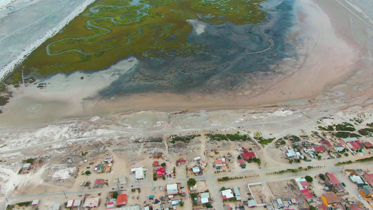 Drone view of green tidal channels above a sandy settlement with red-roofed houses.