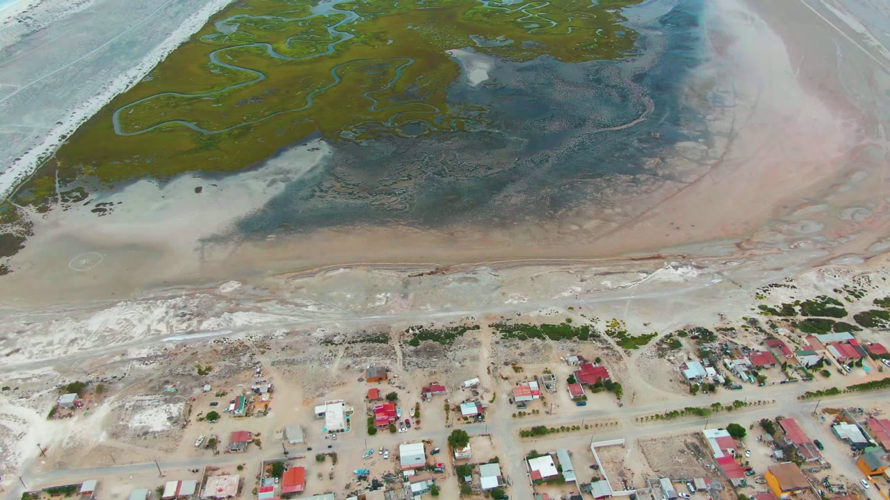 Aerial view in Punta Abreojos showing pale surf along the left edge, green marsh channels across the top, pink-toned tidal flats to the right, and clustered shore buildings below, with no visible people.