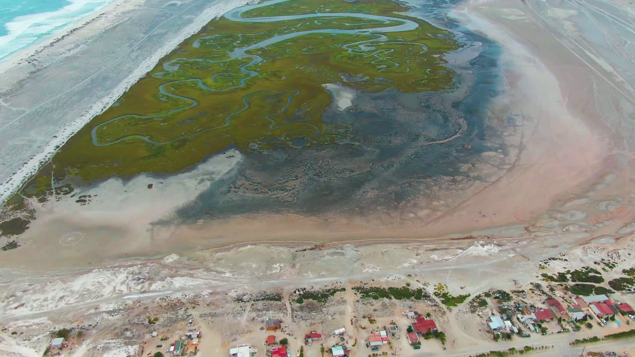 Aerial frame over Punta Abreojos showing green wetland channels, dark mudflats, pale shoreline, turquoise water, and red-roofed shore houses at the bottom with no visible people.