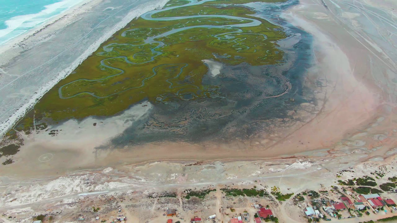 Punta Abreojos aerial frame showing the wetland island and channel network above dark mudflats, with pale sand and red-roofed houses still visible along the bottom edge and no visible people.