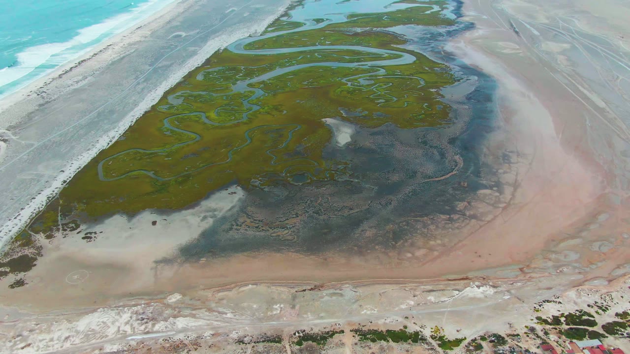 High aerial frame over Punta Abreojos, Baja California Sur, showing turquoise surf at left, green lagoon channels in the center, pale sandbars, pink-tan wet shoreline, and no visible people.