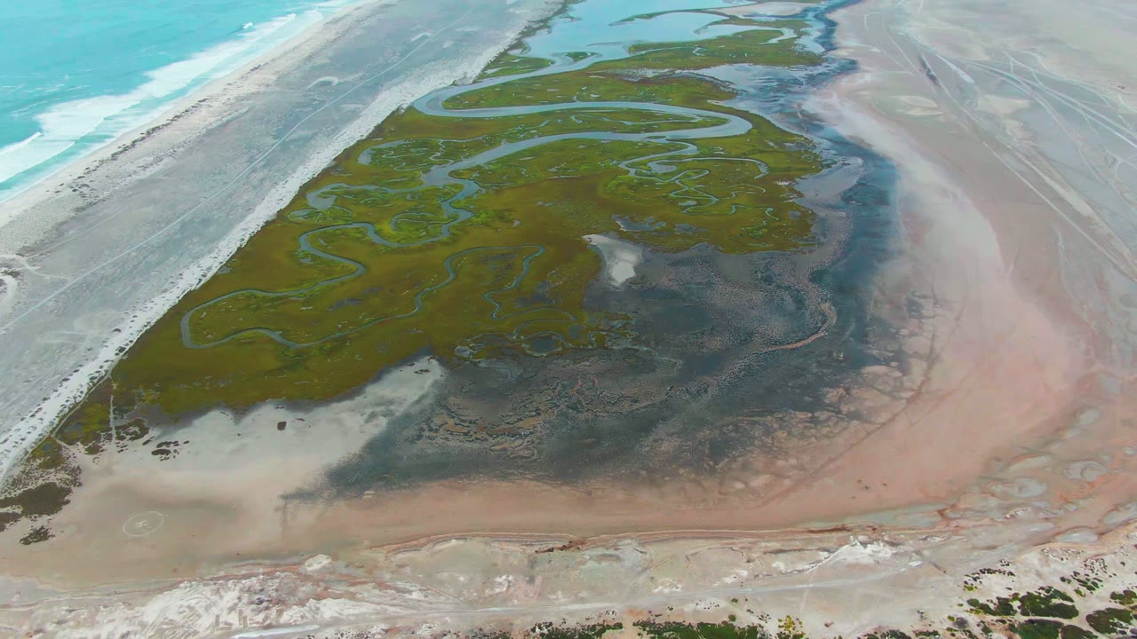 Punta Abreojos aerial frame showing winding blue-green lagoon channels across darker wetland patches, with white surf and beach on the left and no visible people.