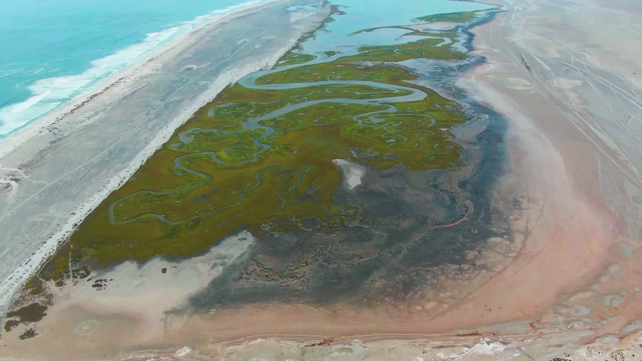 Aerial view over Punta Abreojos, Baja California Sur, with turquoise ocean along the left edge, pale beach, green lagoon flats, winding tidal channels, and no visible people.
