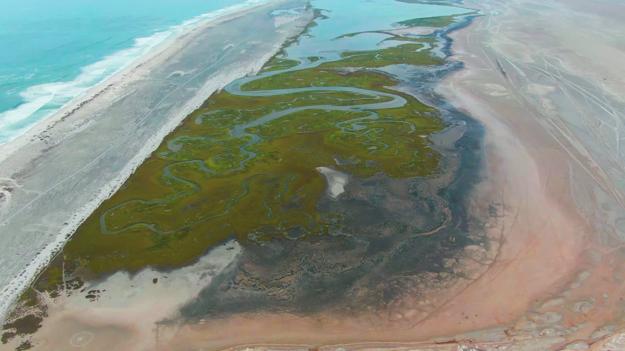 Opening aerial frame over Punta Abreojos, Baja California Sur, with turquoise ocean at left, pale beach, green wetland channels, pink tidal flats, and no visible people.