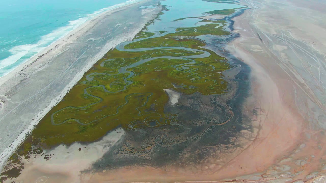 Drone-supported aerial view over Punta Abreojos with the beach and breaking waves on the left, green wetland water paths in the center, and pink-tan flats to the right, with no visible people.