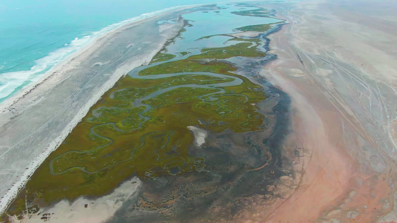 At 0:00, a high aerial view over Punta Abreojos shows green marsh channels running beside a pale sandy shore and turquoise ocean, with no visible people.