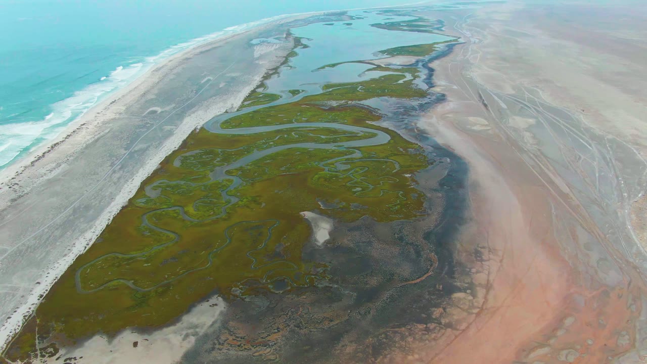 Drone view of winding wetland channels, sand flats, and surf-lined beach.