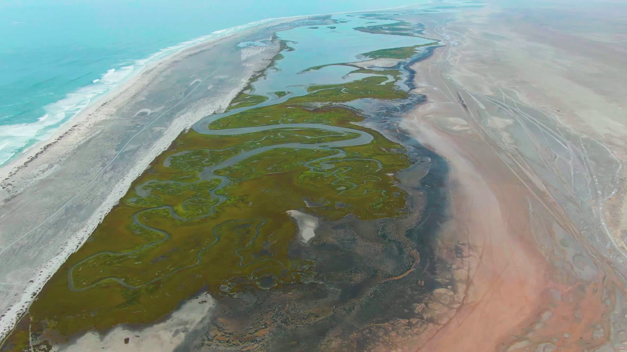 At 0:02, the aerial composition keeps the turquoise ocean on the left and the marsh in the center, with pinkish sand flats and shallow tidal water extending to the right, with no visible people.