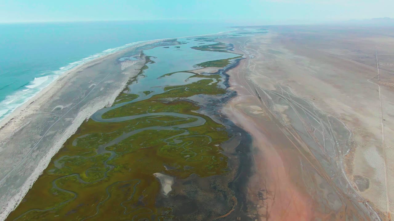 Aerial frame over Punta Abreojos, Mexico, showing turquoise ocean at left, a pale beach strip, green wetland water and channels through the center, dry plain at right, and no visible people.