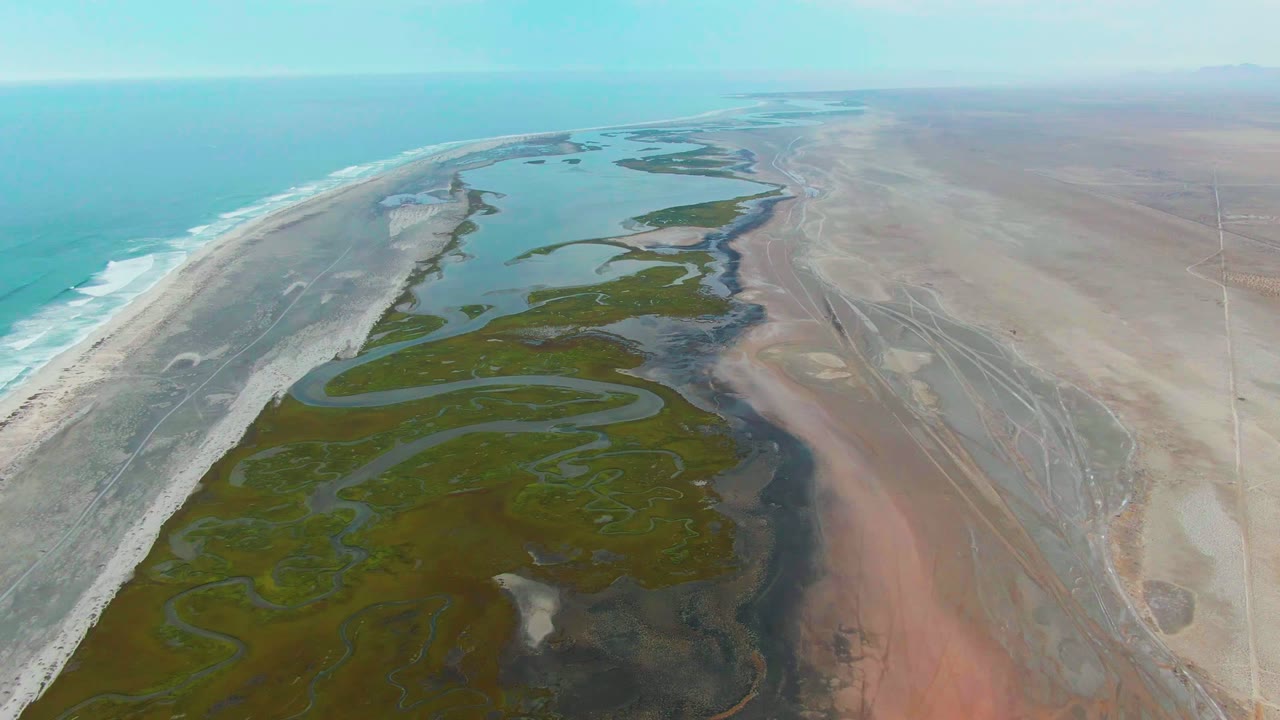 High coastal aerial with surf, beach, marsh pools, and tan desert plain.