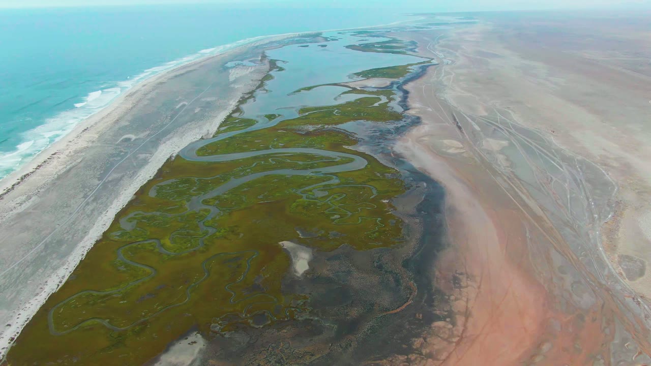 Aerial view over Punta Abreojos, Baja California Sur, with turquoise ocean surf on the left, green tidal wetlands in the center, dry flats on the right, and no visible people.