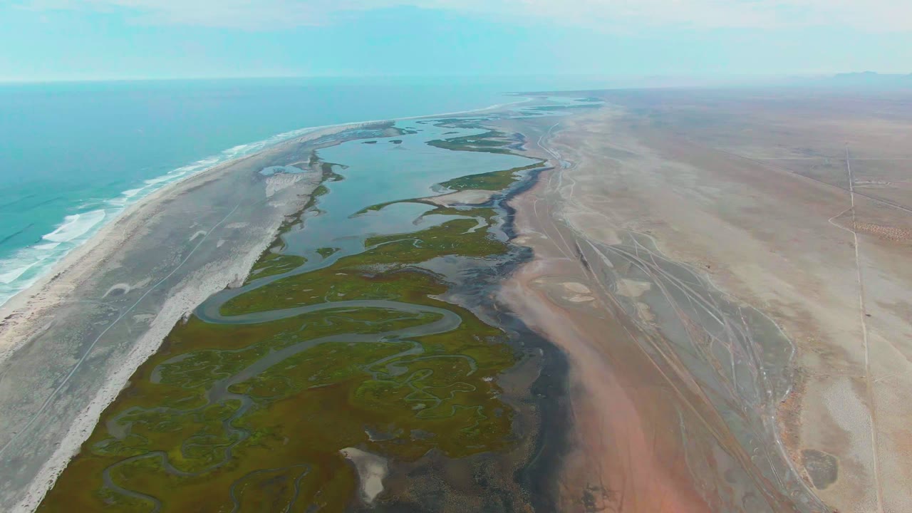 Aerial view over Punta Abreojos with turquoise ocean surf at left, a pale sand barrier, green winding wetland channels through the center, dry brown terrain at right, and no visible people.