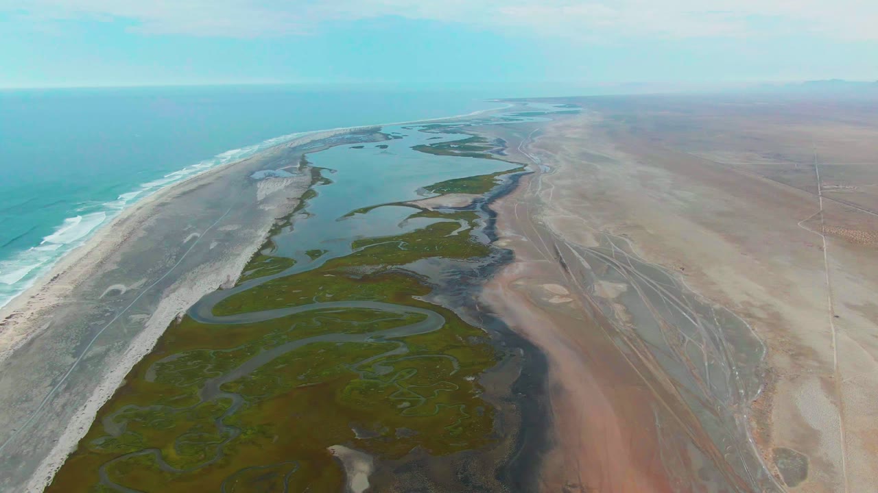 Drone-like aerial over Punta Abreojos with blue-green ocean to the left, white waves meeting the sand, a branching wetland in the middle, brown dry land to the right, and no visible people.