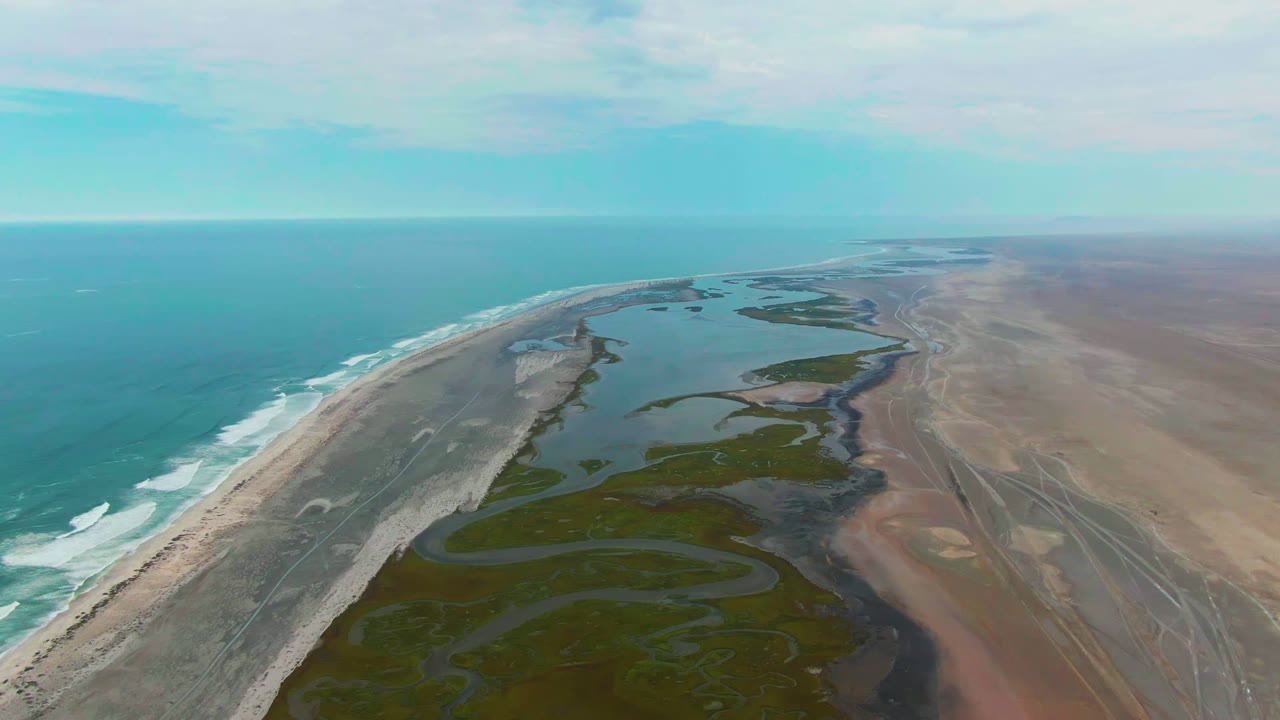 Wide aerial frame over Punta Abreojos, Baja California Sur, with turquoise ocean on the left, a sandy surf edge, green wetland channels through the middle, tan flats on the right, and no visible people.
