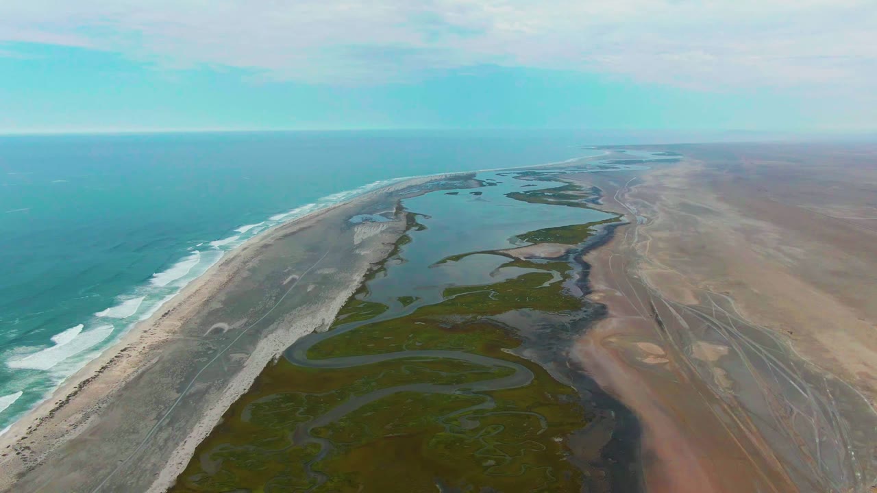 The 0:02 frame holds a high drone perspective over Punta Abreojos, with surf bands along the left edge, green marsh patches and dark channels below center, open lagoon water ahead, and no visible people.
