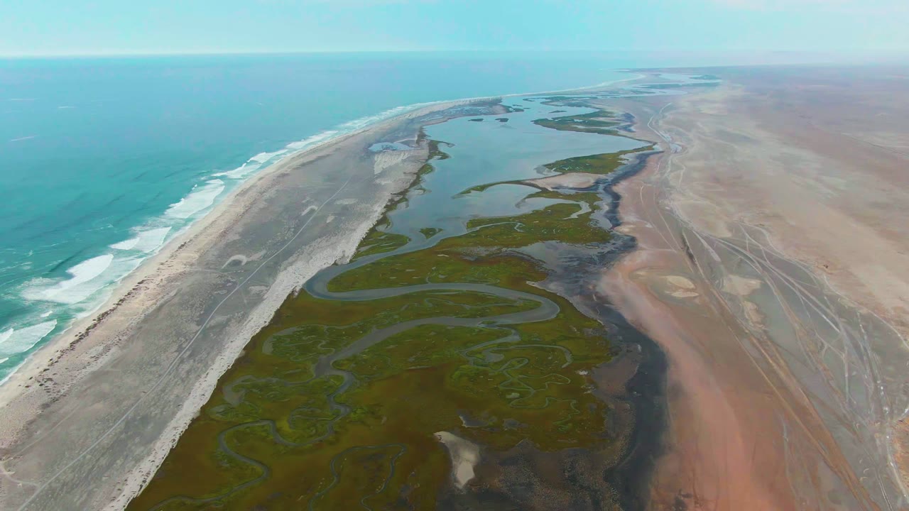 Wide aerial frame over Punta Abreojos, Baja California Sur, showing turquoise ocean on the left, a pale beach, green wetland channels, reddish arid flats, and no visible people.
