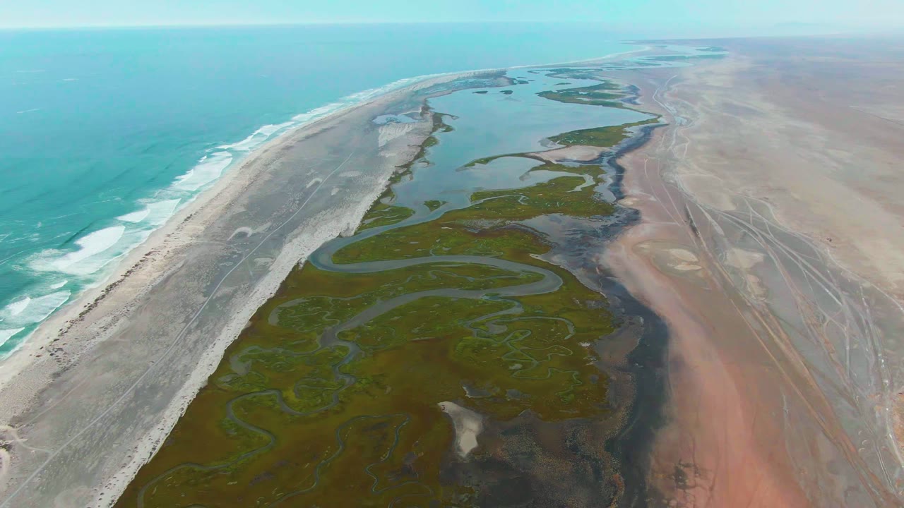 High drone frame over Punta Abreojos showing surf curling along the left edge, a pale sand ridge, irregular green wetland pools, reddish dry flats, and no visible people.