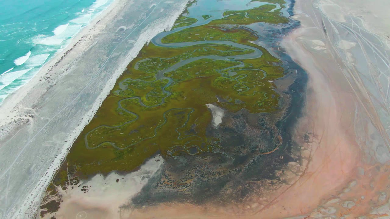At 0:00, an aerial view over Punta Abreojos shows turquoise surf, a pale coastal road, green wetland channels, pink salt flat, shallow pools, and no visible people.
