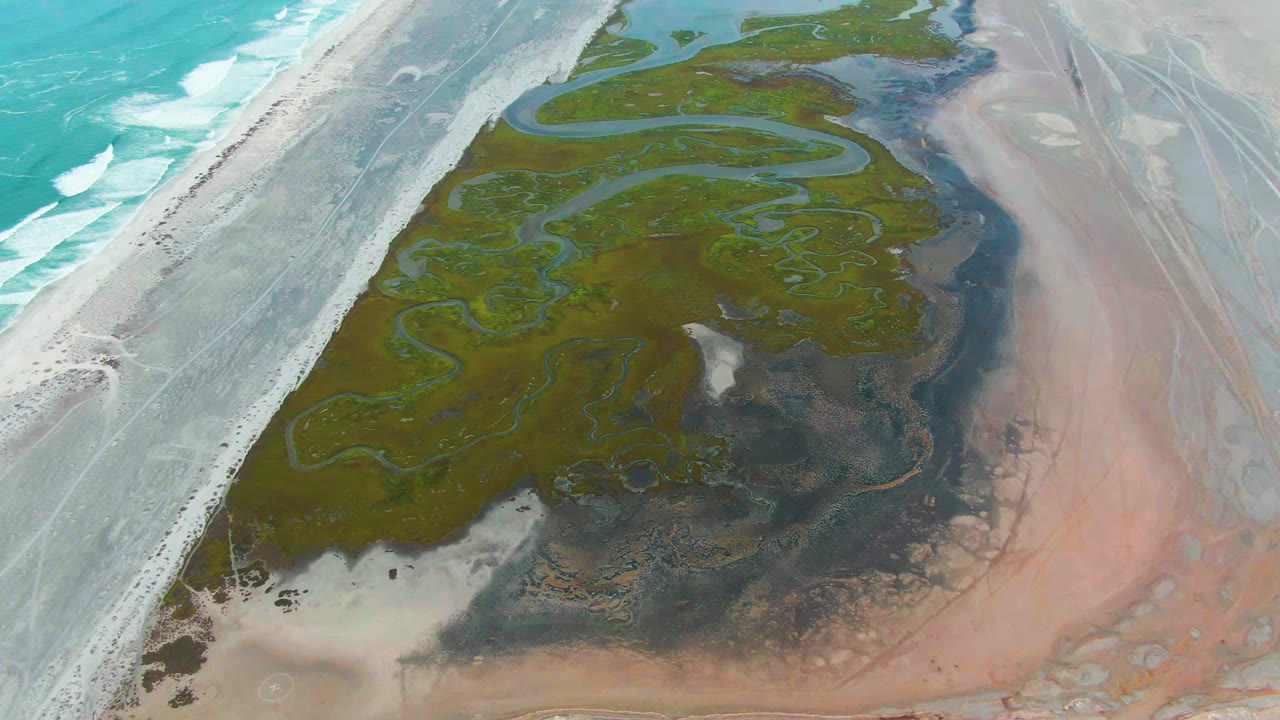 Aerial shoreline view with surf, road, green tidal channels, and exposed pink flats.