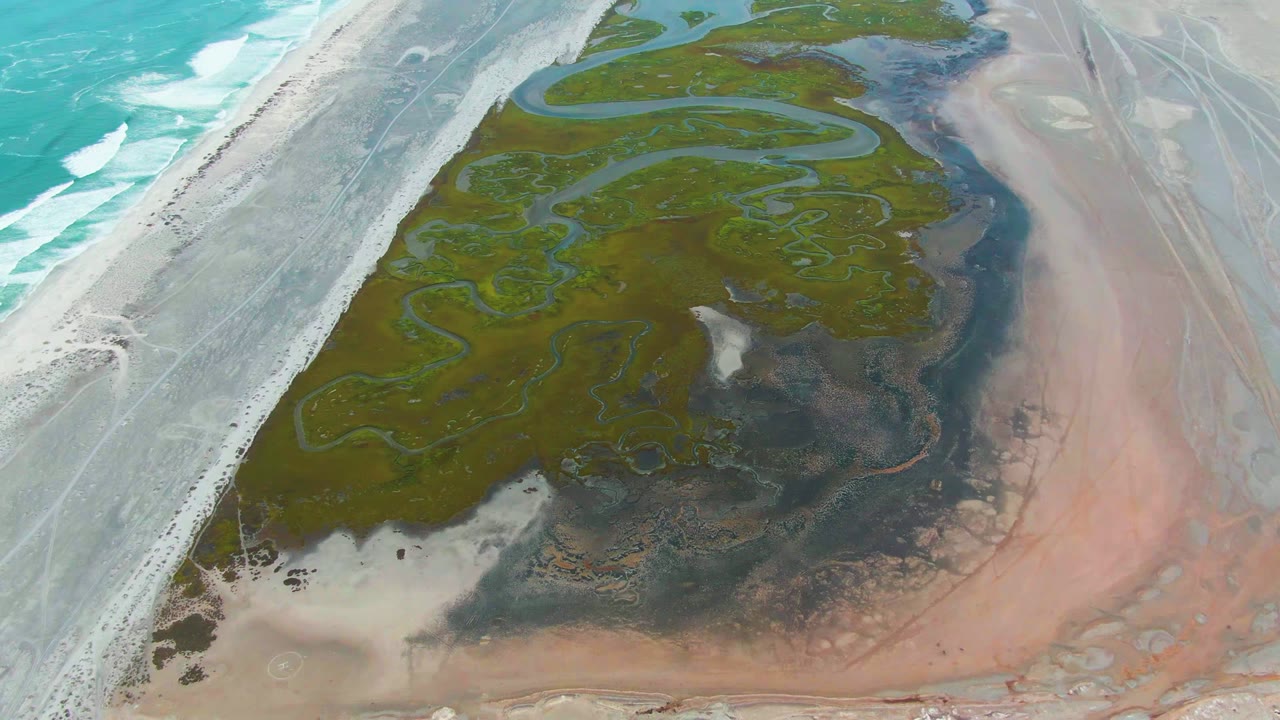 At 0:02, the aerial view over Punta Abreojos shows sinuous green wetland channels bordered by surf and a pale road on the left and pink salt flat on the right, with no visible people.
