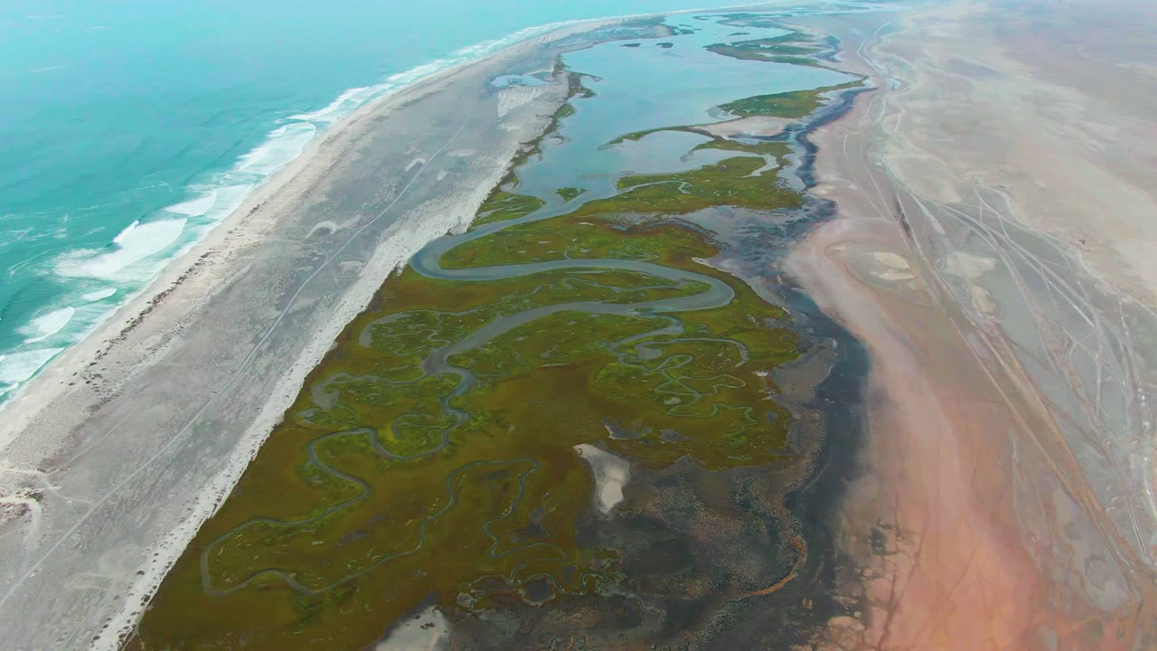 Aerial view at Punta Abreojos, Baja California Sur, showing turquoise surf on the left, a pale sand strip, green tidal channels, pink-tan flats, and no visible people.