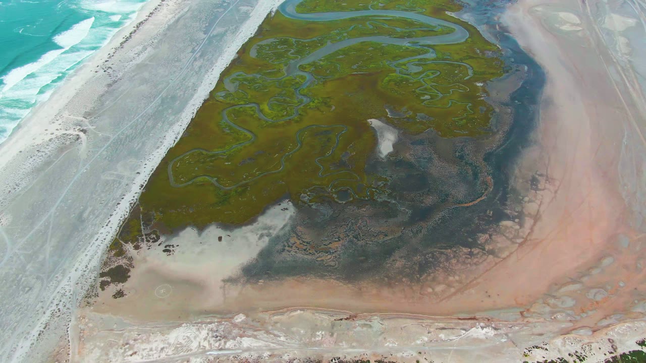Aerial frame over Punta Abreojos, Baja California Sur showing turquoise surf at left, a pale causeway, green algae channels, dark mudflat, pink shallow lagoon water, and no visible people.