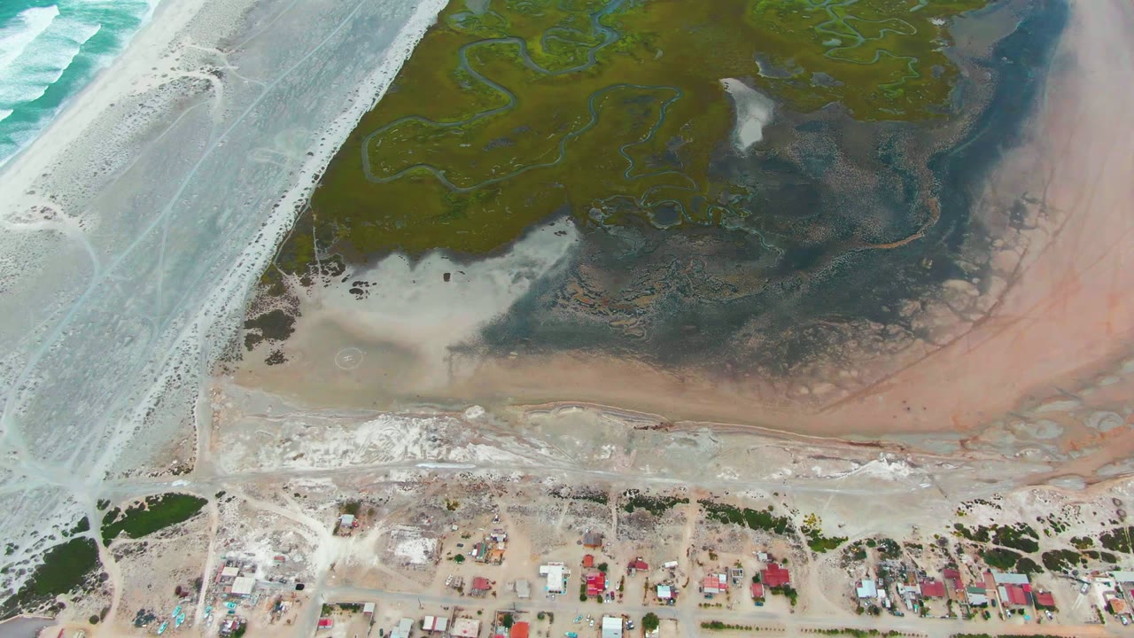 Opening aerial frame over Punta Abreojos, Baja California Sur, showing pale surf at left, green wetland channels above, exposed mudflats near shore, small houses below, and no visible people.